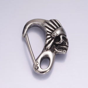 May include: Silver-toned metal key chain featuring a detailed skull design wearing a headdress. The skull has a toothy grin and is attached to a carabiner clip.