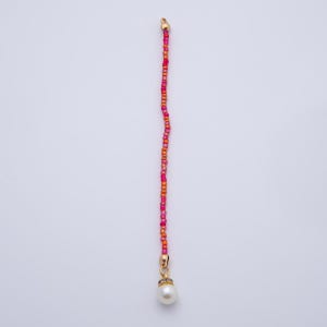 May include: A beaded accessory with a gold-colored clasp and a white pearl pendant. The beads are a mix of pink and orange hues. The accessory is vertically oriented against a white background.