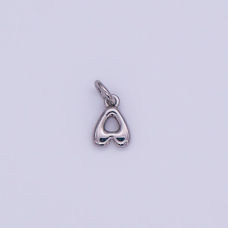 May include: A silver-colored charm in the shape of the letter "A". The charm has a rounded, bubble-like design with a small loop at the top for attaching to a chain or bracelet. The background is a solid, light purple color.
