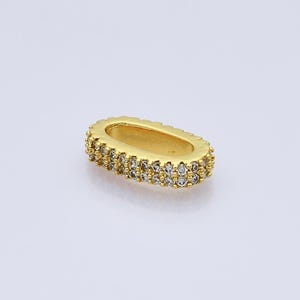 May include: A gold-colored, oval-shaped jewelry component. The piece is encrusted with two rows of small, clear gemstones. The item is set against a plain, light background, highlighting its design and details.