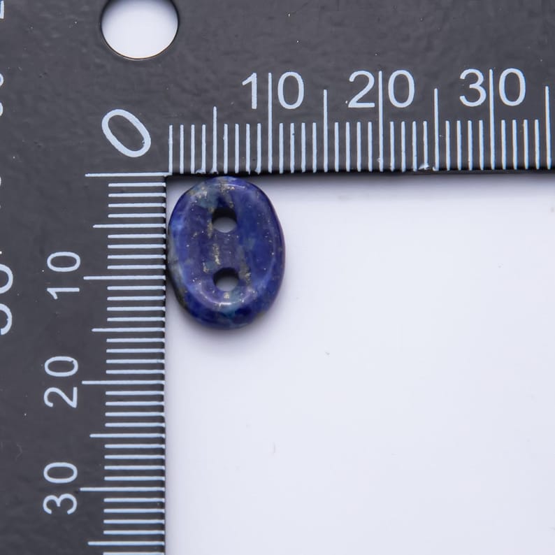 May include: Oval-shaped, dark blue lapis lazuli bead with two holes. The bead is approximately 0.5 inches long. The bead has a polished surface with flecks of gold. The bead is set against a white background and a ruler.