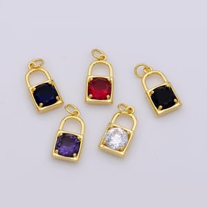 Op de afbeelding: Vijf gouden slotbedels met verschillende gekleurde edelstenen. De bedels hebben de vorm van een hangslot met een ronde edelsteen in het midden. De edelstenen zijn rood, blauw, zwart, paars en helder.