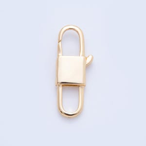 May include: A gold-colored lobster clasp, a jewelry component, is displayed against a white background. The clasp has a rectangular body with a curved top and bottom, and a small lever on the side for opening and closing.
