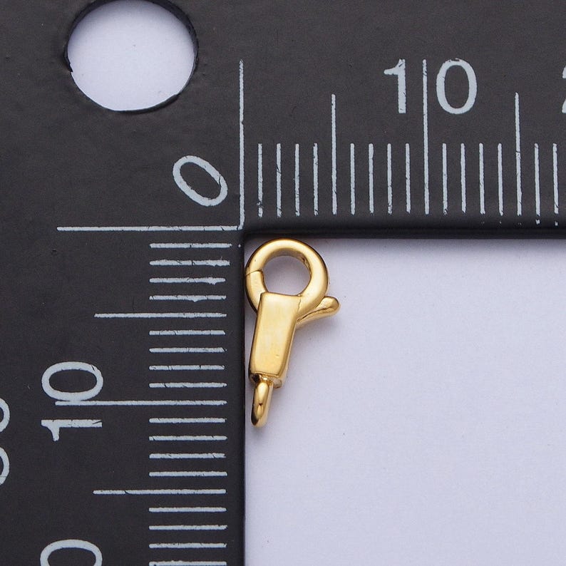 May include: Gold-toned metal lobster clasp with a spring-loaded mechanism. The clasp is approximately 2.5 cm long and is measured against a ruler.
