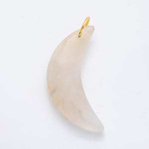 Op de afbeelding: Een maanvormige hanger met een bleke, doorschijnende uitstraling. De hanger heeft een goudkleurige lus aan de bovenkant om op te hangen. Het ontwerp is glad en gebogen, met subtiele kleurvariaties.
