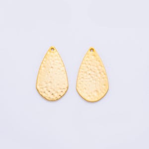 May include: Two gold-colored, teardrop-shaped earring pendants with a hammered texture. Each pendant has a small hole at the top for attachment. The pendants are displayed against a white background.
