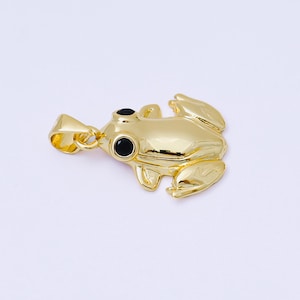May include: A gold-colored frog pendant with black eyes. The pendant is detailed with a textured surface and a loop for a chain. The frog is in a relaxed pose, with its legs extended.