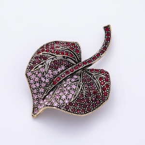 May include: A leaf-shaped brooch featuring a metallic gold-tone frame filled with small, round, faceted crystals. The design displays a color gradient transitioning from deep red at the edges to lighter shades of pink toward the center.