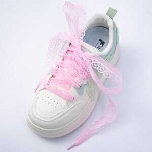 May include: White and pastel green sneaker with pink lace ribbon. The shoe has a white sole and a light green pull tab on the back. The laces are replaced with a pink lace ribbon, adding a unique touch to the footwear.