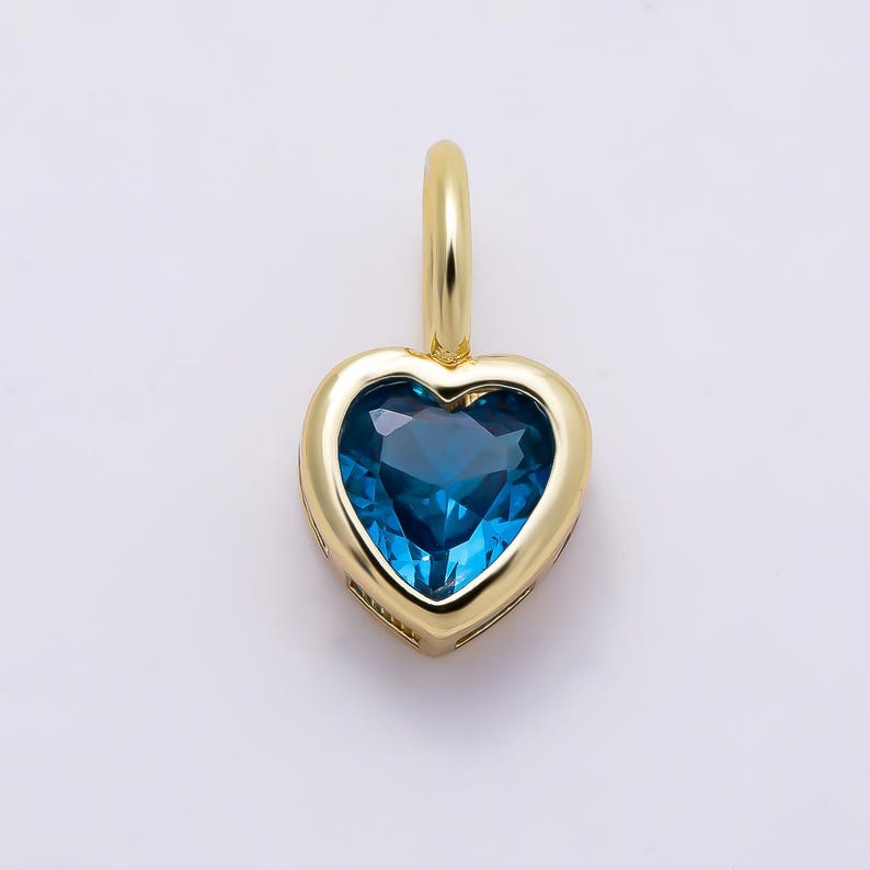 May include: A heart-shaped pendant featuring a deep blue gemstone set in a polished yellow gold frame. The pendant includes a small, smooth gold bail at the top for attachment to a chain, showcasing a clean, symmetrical design with a vibrant blue center.