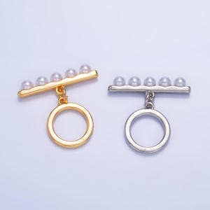 Op de afbeelding: Twee gouden en zilveren toggle-sluitingen met een rij van vijf nepparels en een hangende ring.