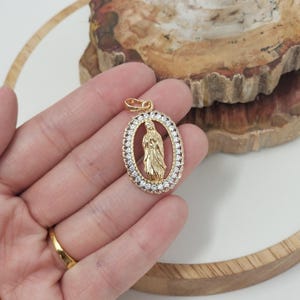 Puede incluir: Pendiente ovalado de oro con un borde de cristal transparente y una representaci&oacute;n de la Virgen Mar&iacute;a en su interior.