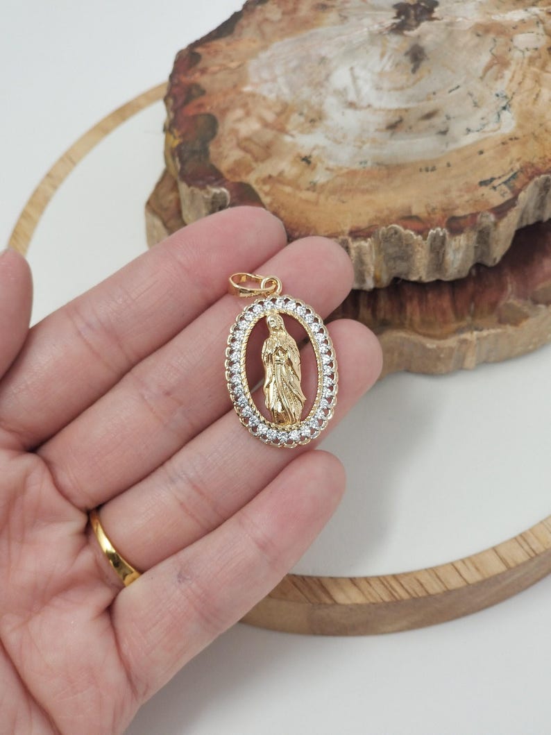 Puede incluir: Pendiente ovalado de oro con un borde de cristal transparente y una representaci&oacute;n de la Virgen Mar&iacute;a en su interior.