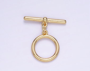 Puede incluir: Un cierre de palanca dorado, que presenta una barra horizontal y un anillo circular conectados por un pequeño lazo. El cierre está diseñado para la fabricación de joyas y tiene una superficie brillante y reflectante. El fondo es de un color suave y neutro.