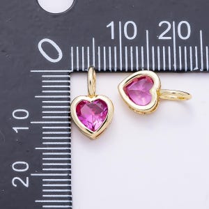 May include: Two gold-toned heart-shaped charms featuring faceted pink stones, each with a small jump ring attached. The charms are positioned next to a black ruler with white markings, showing the items are approximately 0.5 inches in width.