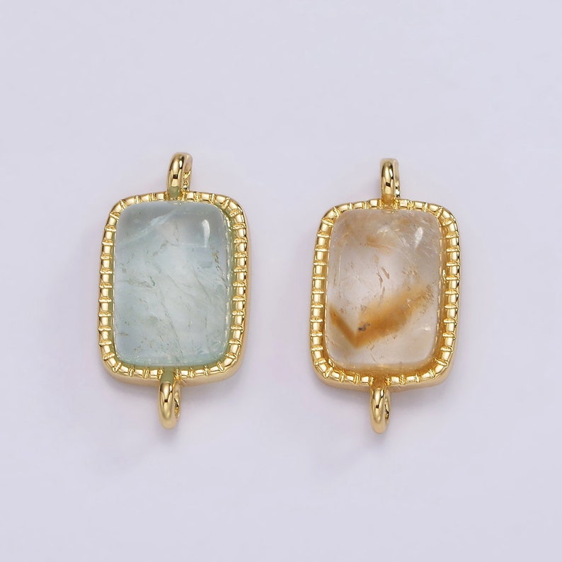 May include: Two gold-toned pendant charms with a rectangular shape. The charms feature a clear stone with a gold-toned border. One stone is light blue and the other is clear with a brown tint.