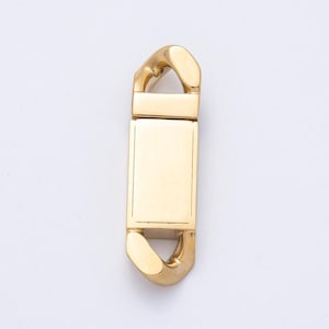 Puede incluir: Un cierre rectangular dorado con extremos redondeados y una placa rectangular central. El cierre tiene un acabado pulido y está sobre un fondo blanco. El diseño es simple y elegante.