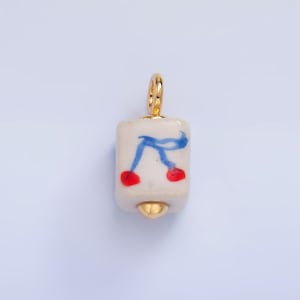 May include: A gold charm with a white ceramic cube featuring a blue and red design. The cube has a gold loop at the top and a gold ball at the bottom.