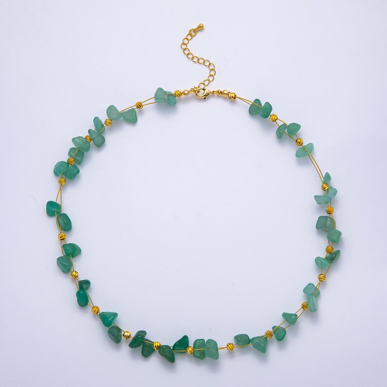 May include: A delicate necklace featuring small, irregular green gemstone beads strung on a thin gold-colored wire. The necklace has a gold-colored clasp and small gold-colored beads interspersed between the green stones. The necklace is designed to be worn around the neck.