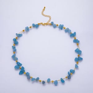 May include: A delicate necklace featuring light blue, irregular-shaped gemstones strung on a gold-colored wire. The necklace has a gold-colored chain clasp and small gold beads interspersed between the gemstones. The necklace is displayed on a white background.