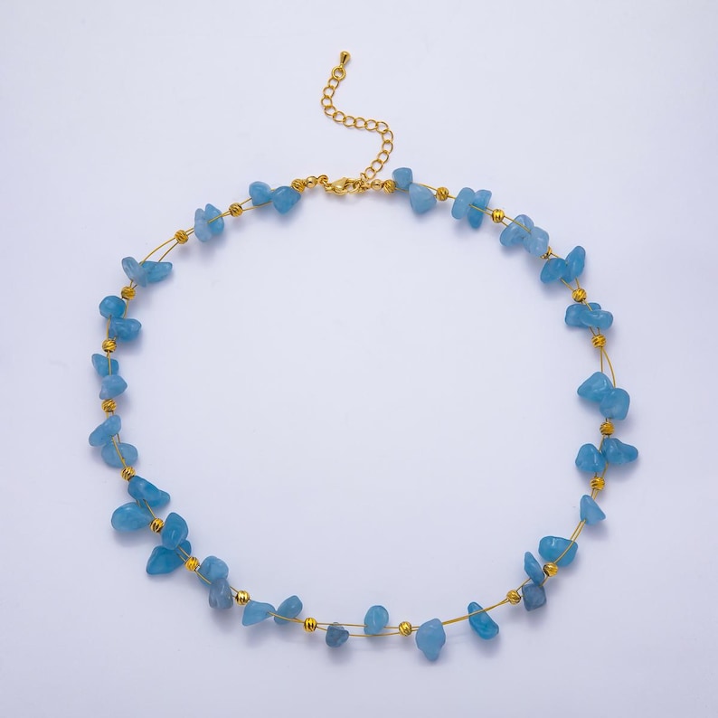 May include: A delicate necklace featuring light blue, irregular-shaped gemstones strung on a gold-colored wire. The necklace has a gold-colored chain clasp and small gold beads interspersed between the gemstones. The necklace is displayed on a white background.