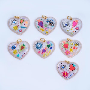 Peut inclure: Sept pendentifs en forme de cœur avec des boucles dorées. Chaque pendentif a un fond blanc avec des motifs peints colorés, notamment des yeux, des cœurs, des fleurs et des formes géométriques. Les pendentifs ont un bord festonné.