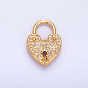 May include: A gold-colored heart-shaped padlock pendant encrusted with small, clear crystals. The pendant has a keyhole and a curved handle. The background is a solid, light color.
