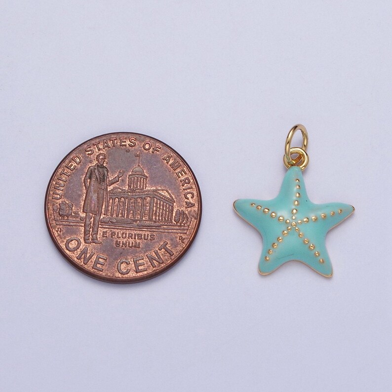 May include: A gold-colored starfish charm with a light blue enamel finish and gold-colored dot accents. The charm is next to a one-cent coin for size comparison. The charm has a small loop for attaching to a necklace or bracelet.