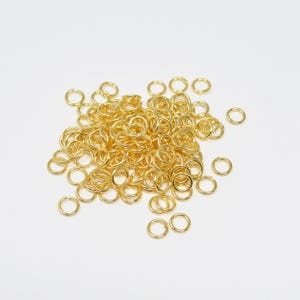 May include: A pile of small, gold-colored jump rings, likely for jewelry making or crafts, are scattered against a white background. The rings are uniform in size and shape, with a bright, metallic finish. They are ready to be used in various creative projects.