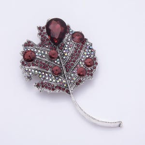 Peut inclure: Une broche en forme de feuille argentée ornée d'un grand cristal rouge foncé en forme de goutte en haut, entouré de cristaux ronds rouge foncé plus petits et de nombreux minuscules strass irisés recouvrant la surface de la structure métallique.