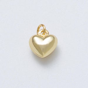 May include: A gold-colored heart-shaped pendant with a rounded, polished surface. The pendant has a small loop at the top for attaching to a chain or bracelet. The heart is a classic symbol of love and affection.
