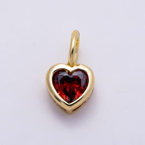 May include: A heart-shaped pendant featuring a deep red gemstone set within a polished gold-tone metal frame. The pendant includes a small, rounded gold-tone bail at the top, designed for attachment to a chain or cord, showcasing a smooth finish.
