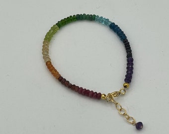 May include: A rainbow gemstone bracelet with a gold clasp and a small amethyst charm. The bracelet is made of small, faceted gemstones in shades of green, blue, purple, and red.