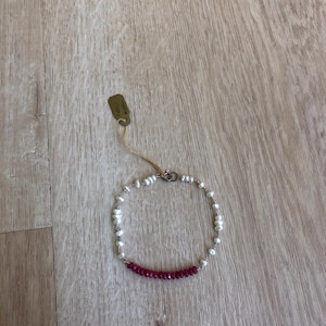 Puede incluir: Un brazalete delicado con una fila de pequeñas perlas blancas y una fila de piedras preciosas de color rojo oscuro. El brazalete está hecho de plata y tiene un cierre.