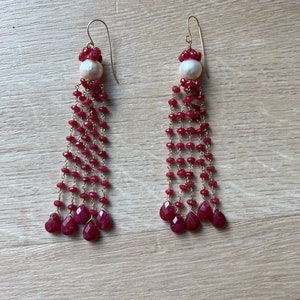 Puede incluir: Un par de pendientes largos y colgantes con una perla blanca en la parte superior y una cascada de cuentas de piedras preciosas rojas. Los pendientes están hechos de oro.