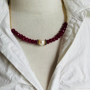 May include: A necklace featuring a strand of deep red, faceted beads, a large white pearl, and a delicate gold chain. The pearl is accented with small, dark embellishments. The necklace is displayed against a white shirt.