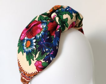 May include: A floral patterned headband featuring a knotted design. The headband displays a cream background with a vibrant pattern of red, pink, blue, and orange flowers and green leaves. The headband is displayed on a white mannequin head.