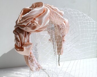 May include: A blush-colored satin headband with a gathered floral design and a delicate white veil. The headband features small, scattered embellishments and a thin, silver-toned band.