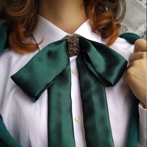May include: A dark green satin bow tie with a beaded embellishment in the center. The bow tie is tied around a white collared shirt.