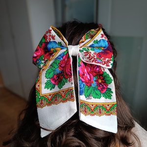 May include: A white bow with a floral pattern in shades of red, pink, blue, green, and yellow. The bow is tied in a large knot and is made of a soft, lightweight fabric.