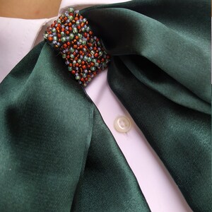 May include: A dark green satin scarf with a beaded embellishment in shades of green, orange, and brown. The scarf is tied around the collar of a white shirt.