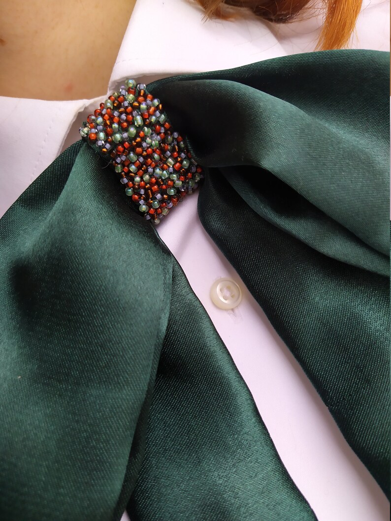 May include: A dark green satin scarf with a beaded embellishment in shades of green, orange, and brown. The scarf is tied around the collar of a white shirt.
