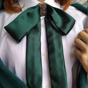 May include: A dark green satin bow tie with a beaded embellishment in the center. The bow tie is tied around the collar of a white button-down shirt.