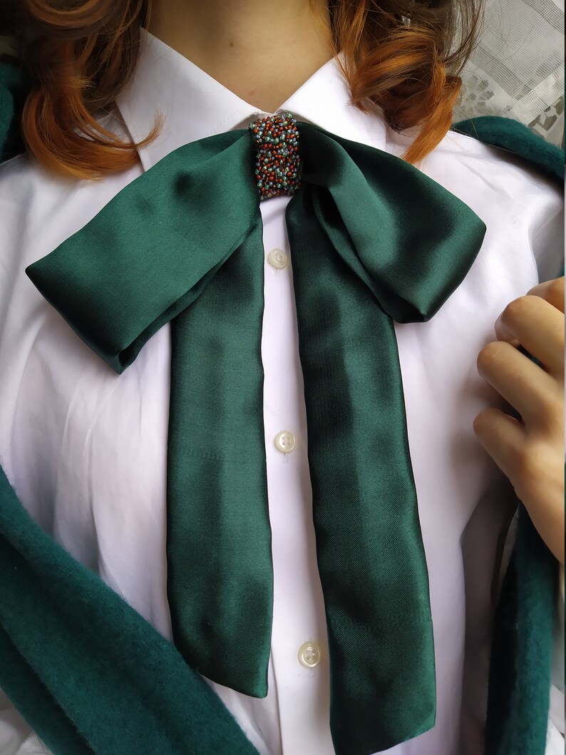 May include: A dark green satin bow tie with a beaded embellishment in the center. The bow tie is tied around the collar of a white button-down shirt.
