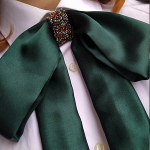 May include: A dark green satin bow tie with a beaded embellishment in the center. The bow tie is tied around a white collared shirt.