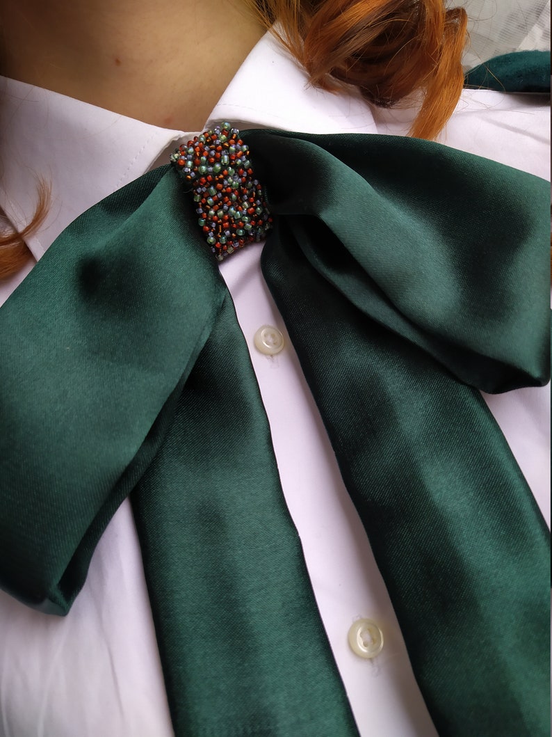 May include: A dark green satin bow tie with a beaded embellishment in the center. The bow tie is tied around a white collared shirt.