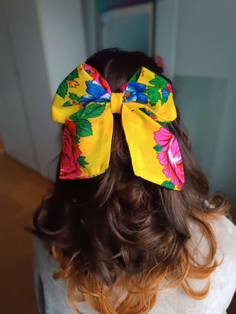 May include: A yellow bow with a floral pattern in pink, blue, and green. The bow is tied at the back of a person's hair.