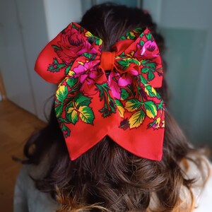 May include: A red bow with a floral pattern in shades of pink, green, and yellow. The bow is tied in a large knot and is worn in the hair.