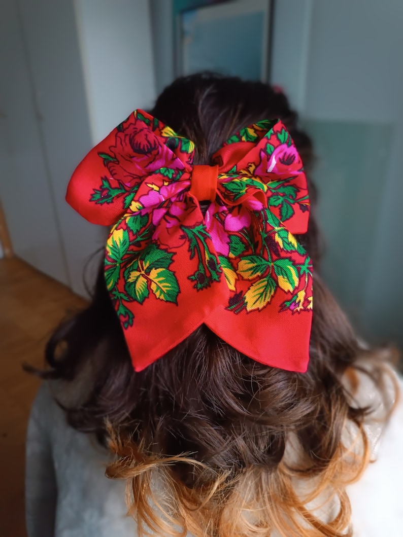 May include: A red bow with a floral pattern in shades of pink, green, and yellow. The bow is tied in a large knot and is worn in the hair.
