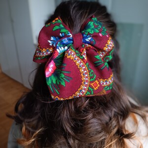 May include: A large burgundy bow with a floral pattern in shades of green, blue, red, and yellow. The bow is tied in a large, full knot and is worn in the hair.